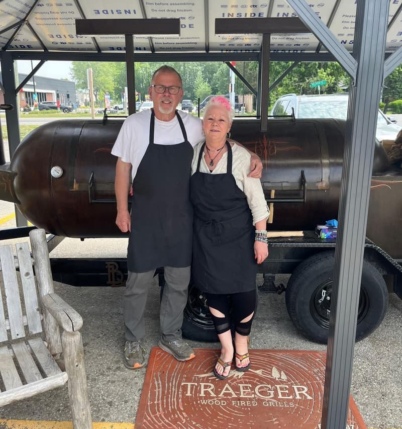 Dar and Papa Jim in black aprons, arm in arm in front of the big black smoker trailer under the canopy outside the store.