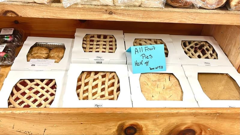 Lattice-top fruit pies in white boxes on a pine shelf, with a handwritten sale sign.