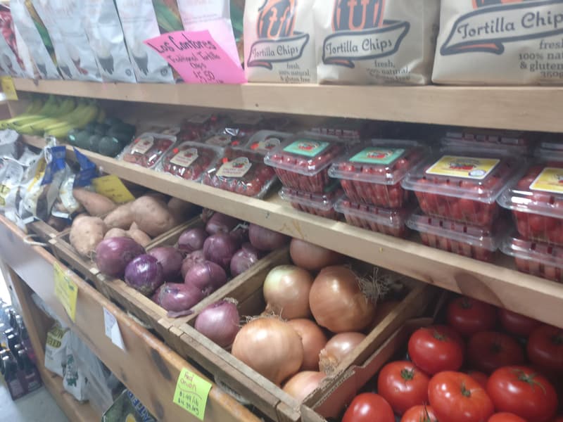 Wooden produce shelves with red onions, cooking onions, sweet potatoes, field tomatoes and pints of strawberries.