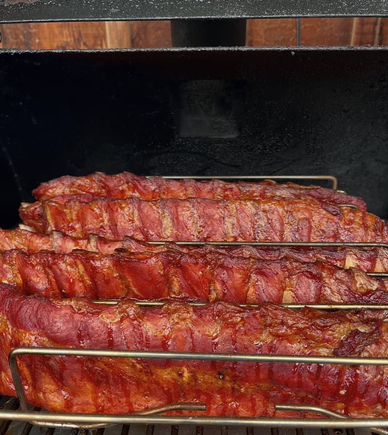 Racks of baby back ribs glazed deep red, stacked on the wire racks inside Dar’s smoker.