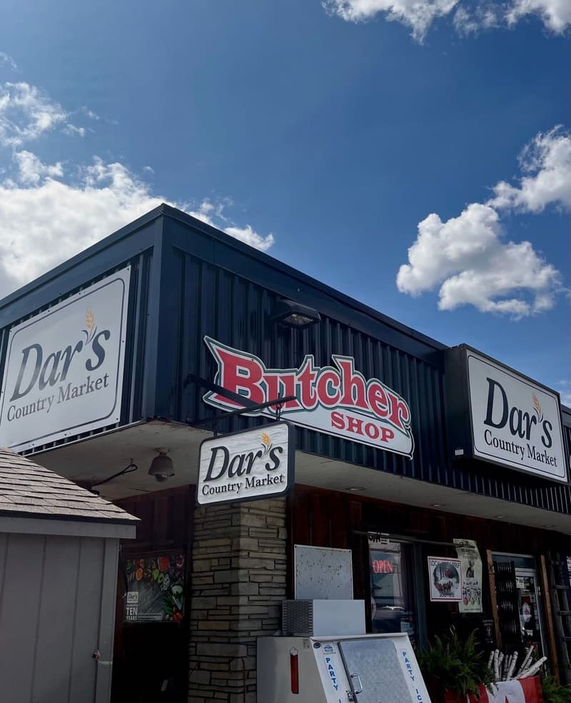 The corner of the store under a blue summer sky, with the red Butcher Shop sign and two Dar’s Country Market signs.